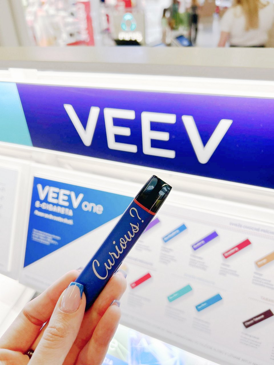 Ruka drží fialové zariadenie VEEV One s nápisom „Curious?“ pred osvetleným predajným stojanom VEEV s prehľadom príchutí v pozadí.