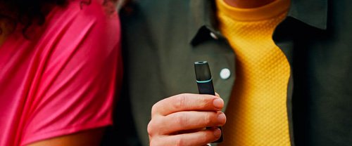 Detail ruky držiacej e-cigaretu tmavej farby so zeleným detailom, v pozadí časť 2 postáv. Jedna v žlto zelenom a druhá v ružovom oblečení.​