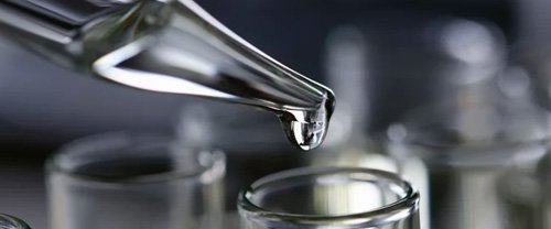Detail kvapkadla s kvapkou tekutiny nad sklenenou nádobou v laboratórnom prostredí.​