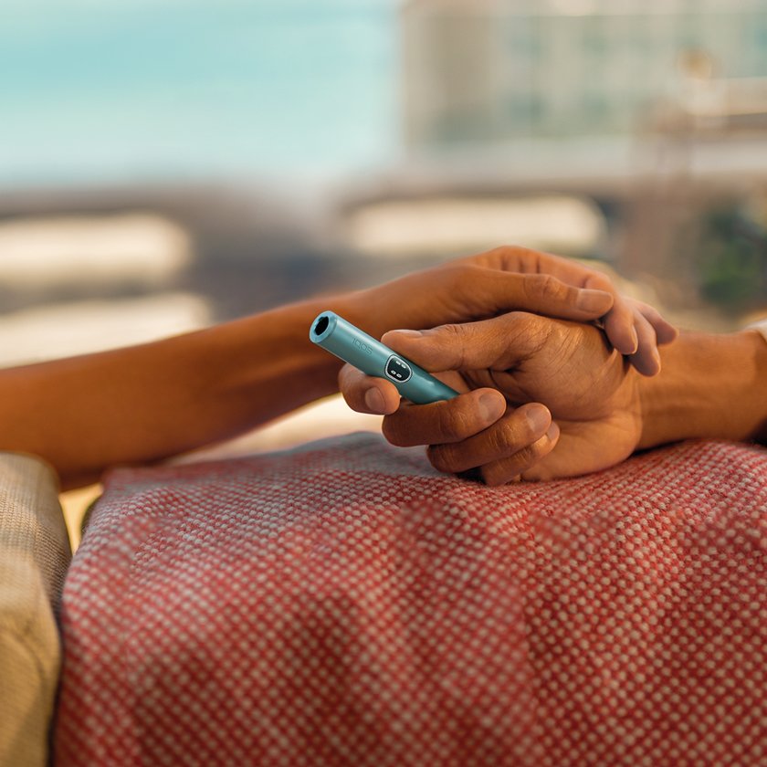 Two hands holding each other, while the one hand is holding a Breeze Blue IQOS ILUMA i device.