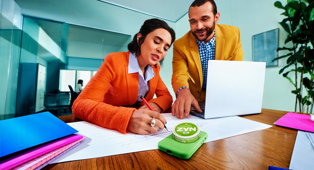Two people working together at a desk with a laptop and papers, with a green ZYN nicotine pouch can resting on a green phone case in the foreground.