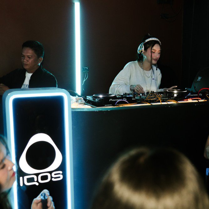 DJ performing live at a nightlife event with professional turntables and prominent IQOS brand signage, while attendees capture the moment on their phones.