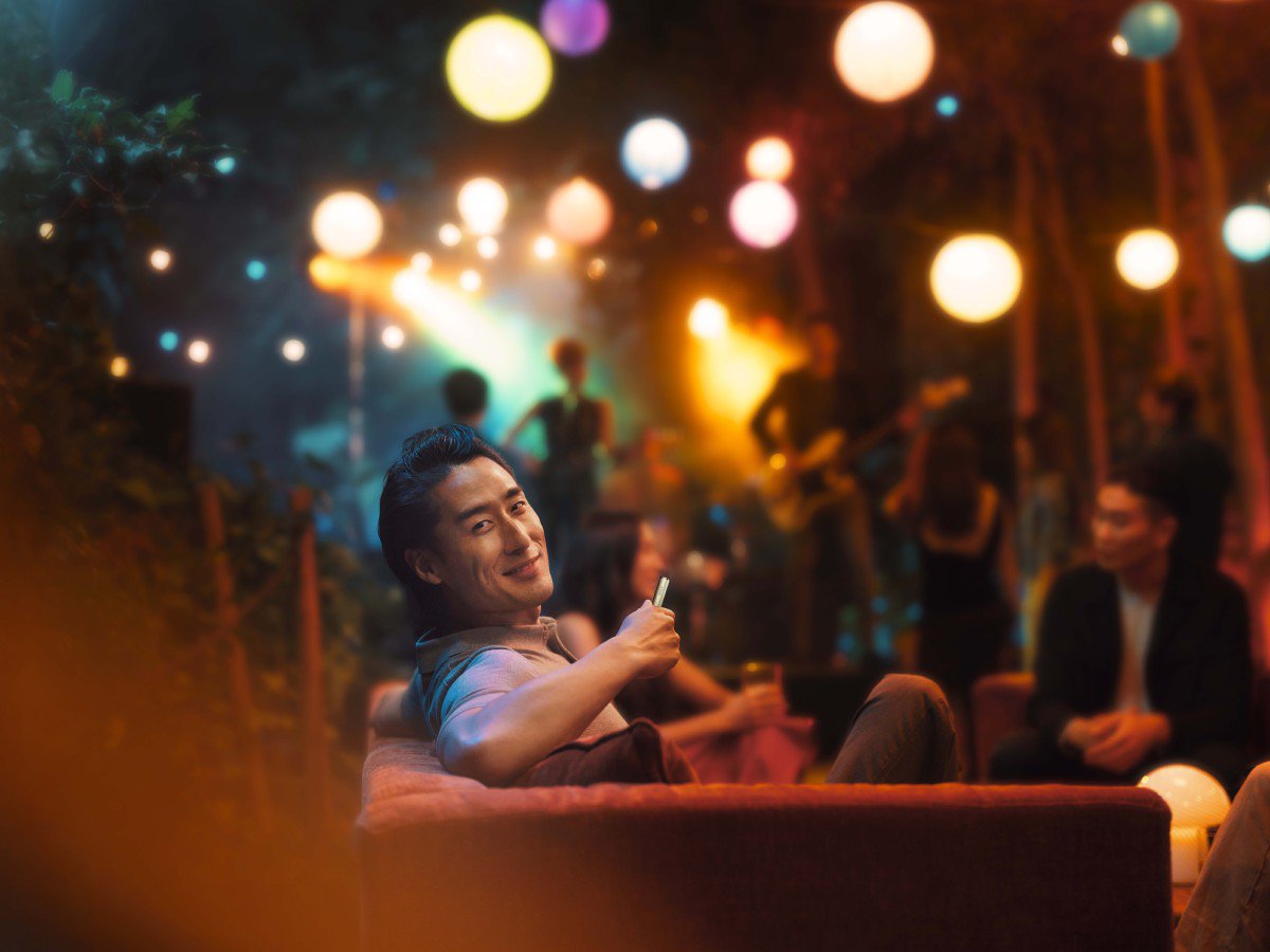A person is sitting on a couch in a dimly lit room with colorful lights and hanging lanterns. There are several people in the background. The atmosphere suggests a relaxed, social setting.