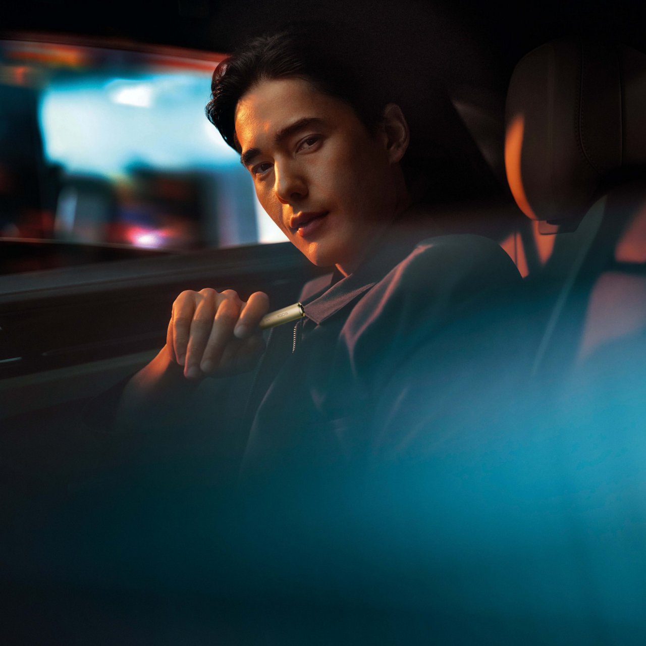 A person is sitting in the passenger’s seat of a car at night. The interior of the car is dimly lit, and there are reflections of lights on the window, creating a blue and orange glow.