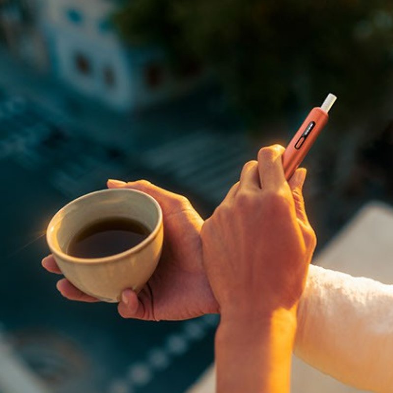 Person hält IQOS Tabakerhitzer und Tasse mit Kaffee in den Händen.