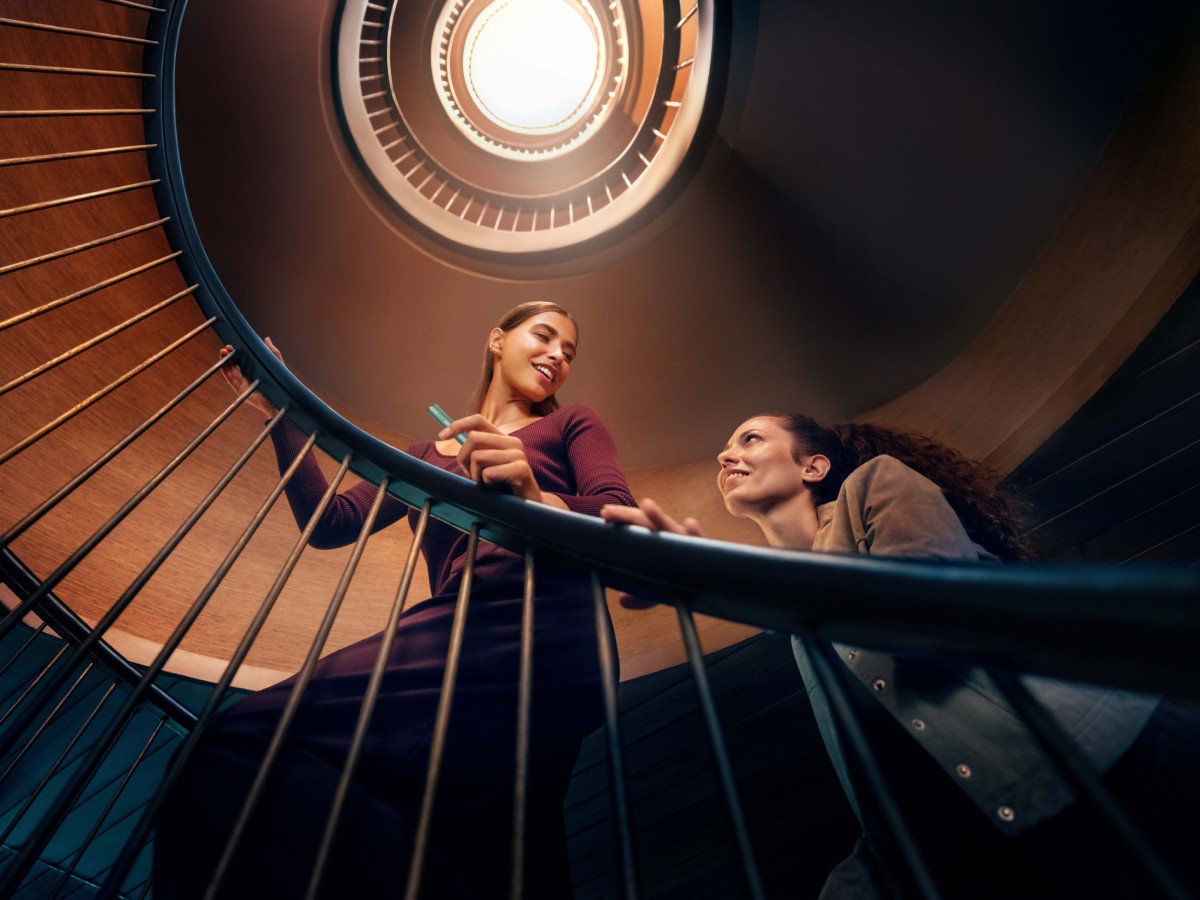 Two people are ascending a spiral staircase. The image is taken from below, looking up towards the ceiling where a circular light is visible. One person is holding onto the railing while the other looks upwards.