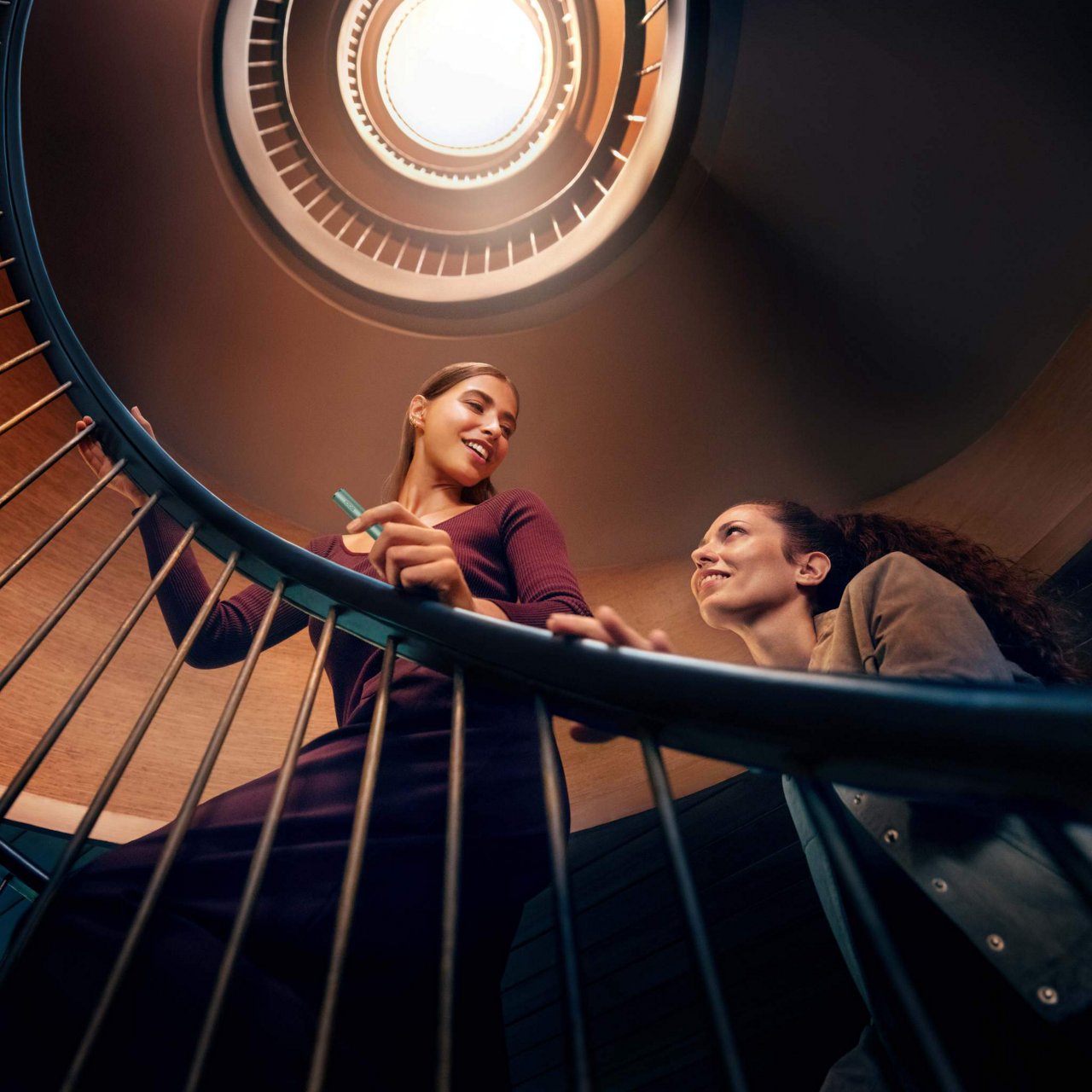 Two people are ascending a spiral staircase. The image is taken from below, looking up towards the ceiling where a circular light is visible. One person is holding onto the railing while the other looks upwards.