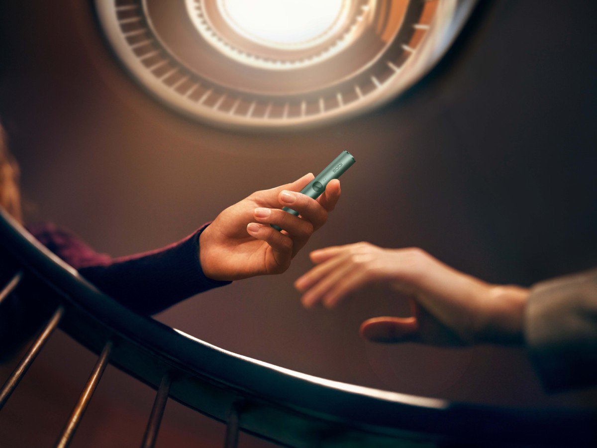A person is handing a Jade Green-colored IQOS ILUMA device to another person. The background features a circular light fixture.