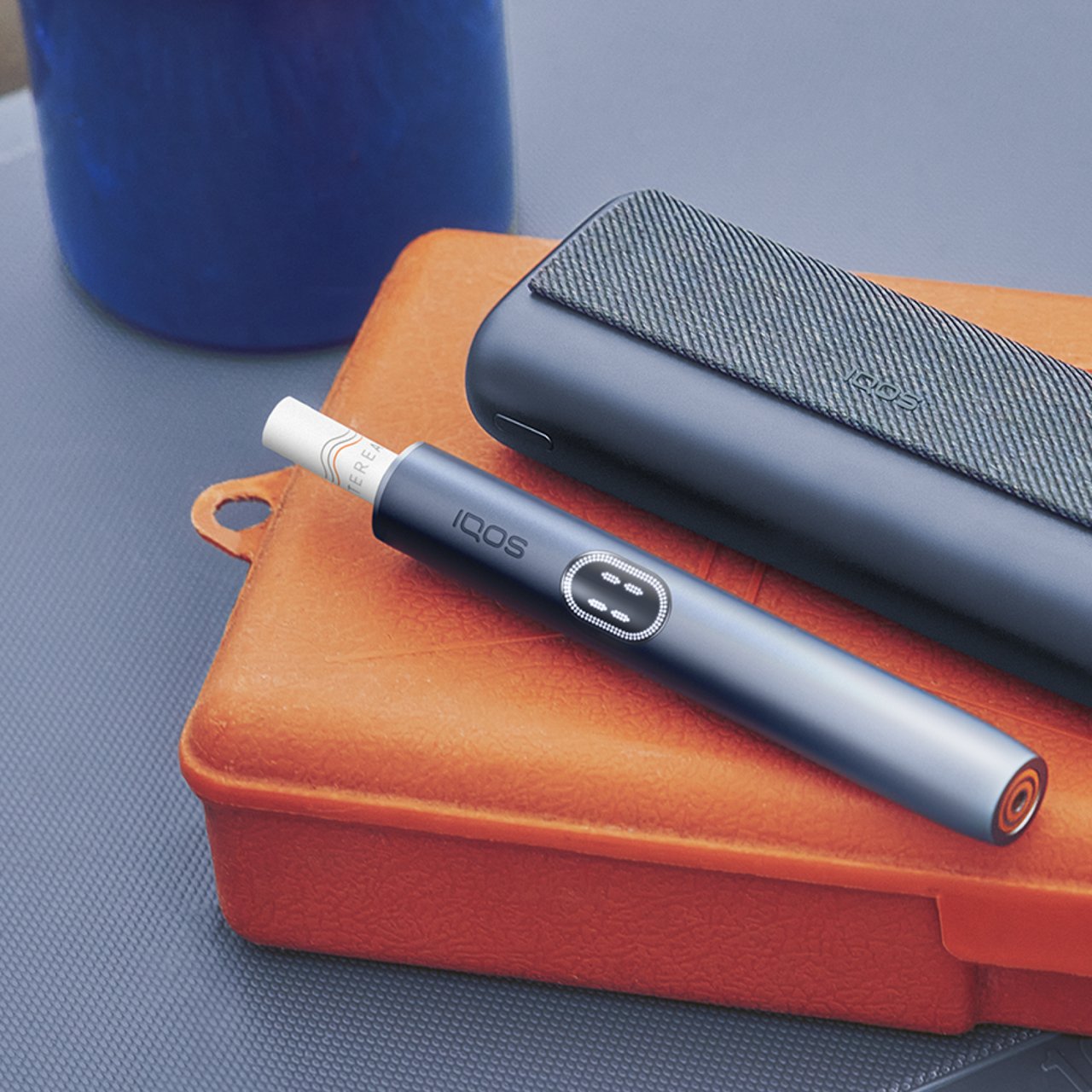 IQOS ILUMA i PRIME Tabakerhitzer liegend auf einer orangefarbenen Dose.