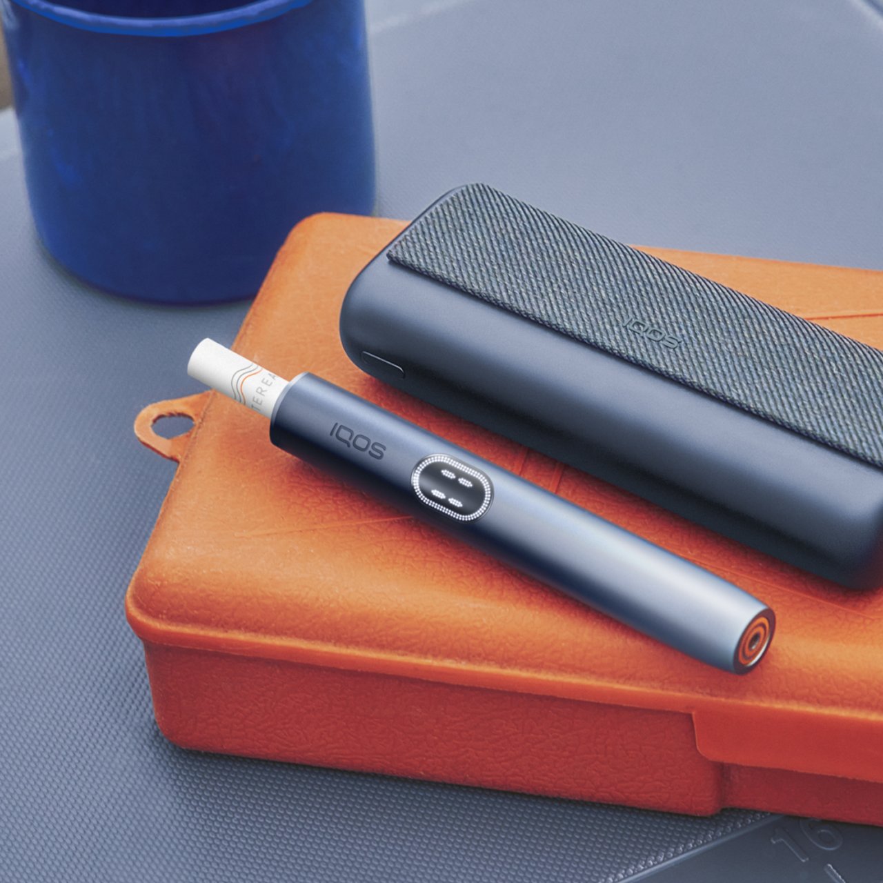  IQOS ILUMA i PRIME Tabakerhitzer liegend  auf einer orangefarbenen Dose.
