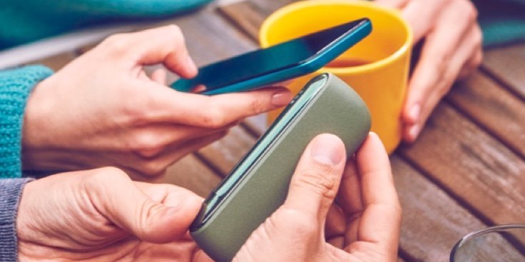 Mehrere Hände an einem Tisch. Sie halten ein IQOS ILUMA, ein Smartphone und eine gelbe Kaffeetasse.