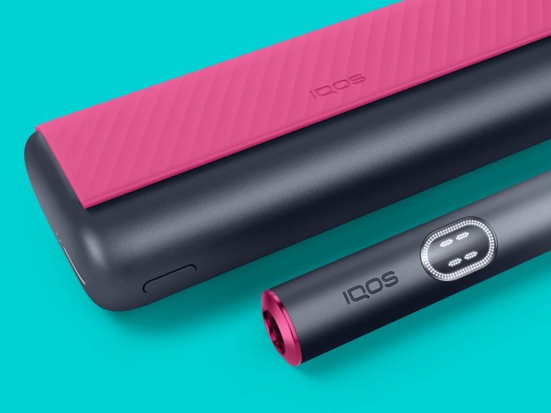 Устройство IQOS ILUMA i PRIME красного и черного цветов и зарядное устройство на светло-бирюзовом фоне