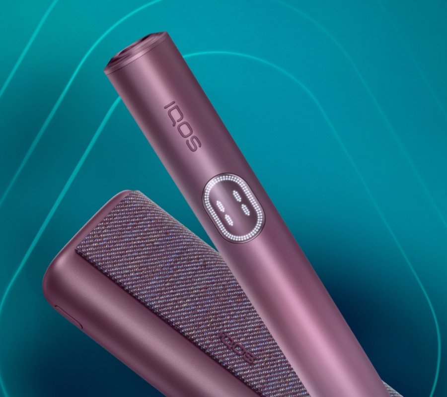 Close-up of IQOS ILUMA i Prime in a metallic plum color with its holder and case displayed against a teal background.
