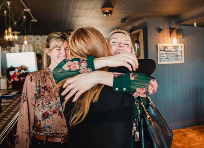 Two women hugging and a third women looking at them and smiling