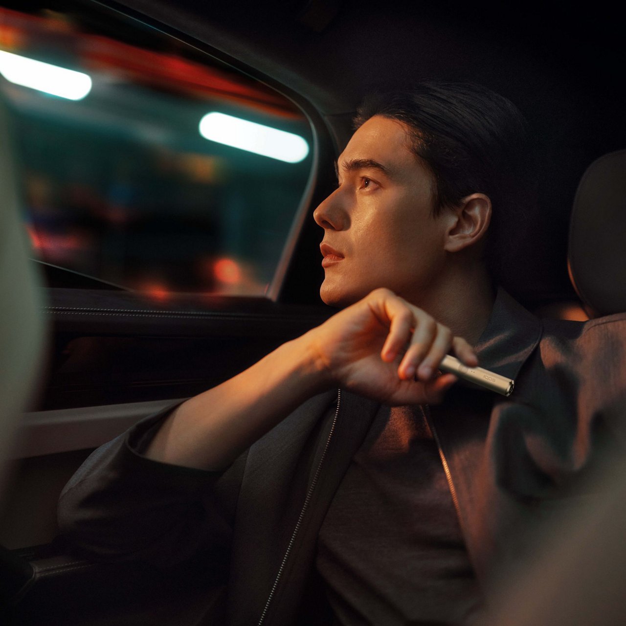 A person sitting in the backseat of a car, looking out of the window. The person is holding an IQOS ILUMA holder in the color Pebble Beige.
