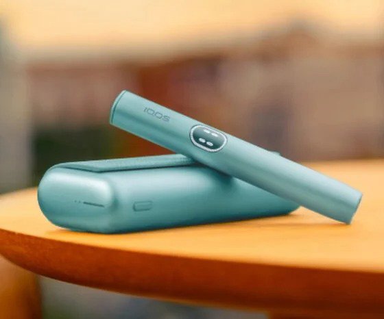 جهاز IQOS ILUMA i Prime باللون الفيروزي مع الحامل والشاحن موضوع على طاولة خشبية بخلفية ضبابية.