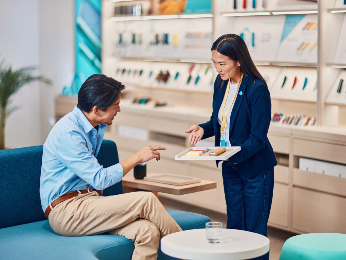 A man sitting on a blue couch in an IQOS store while engaging in conversation with a store sales expert. The sales expert is holding a tray with various items on it. The background shows shelves with neatly arranged products and displays.
