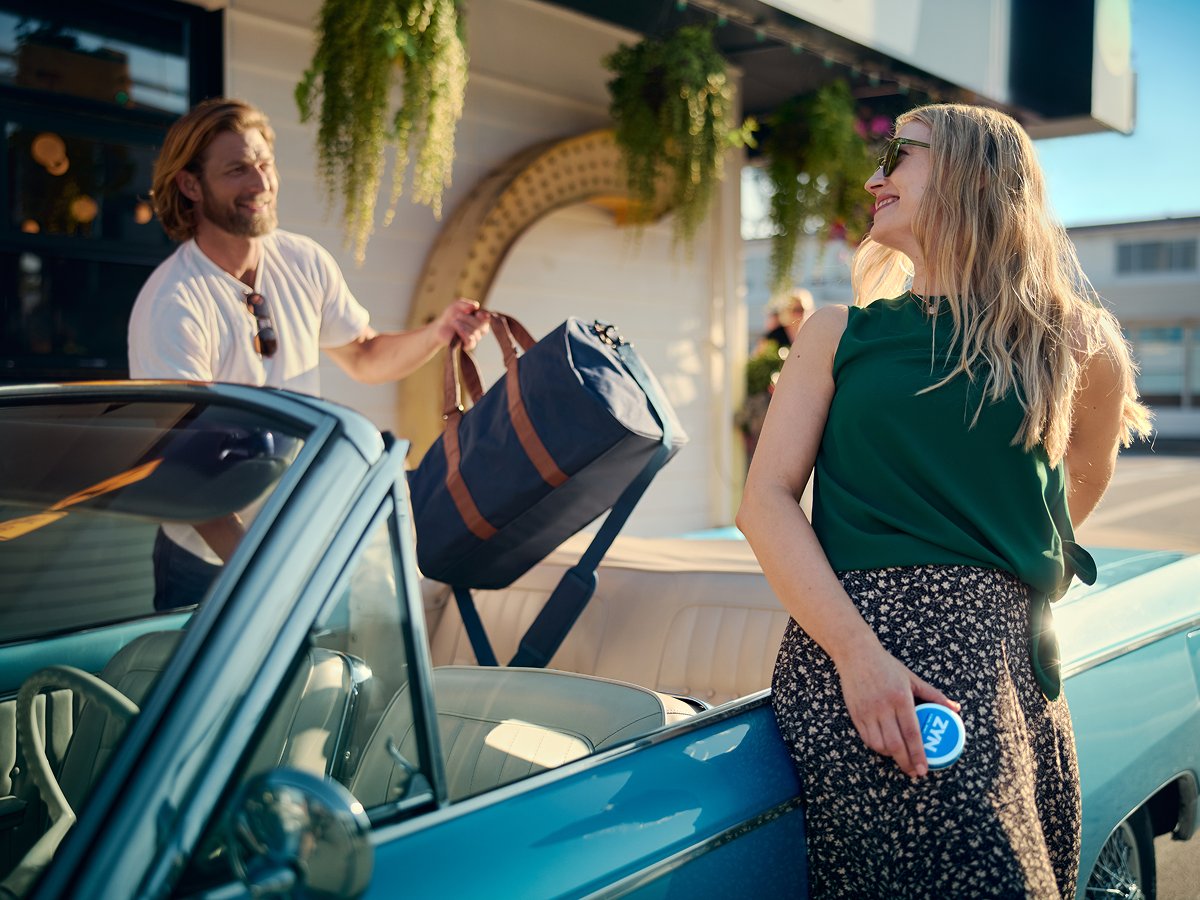 A woman, leaning on the cabriolet, holding a ZYN can and smiling at the guy, putting a bag in the car