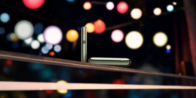 Dos personas con teléfonos móviles sentadas a la mesa; una de ellas también sostiene un dispositivo IQOS ILUMA PRIME color jade blue. 