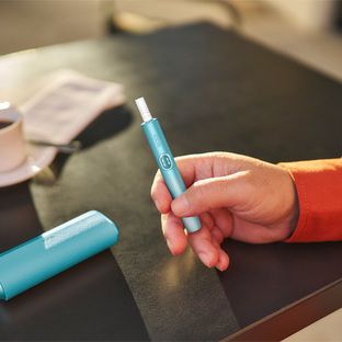 شخصان يحملان هواتف محمولة يجلسان على طاولة؛ يحمل أحدهما أيضًا جهاز IQOS ILUMA Prime باللون الأزرق اليشمي.