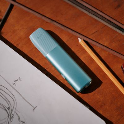 Un IQOS ILUMA i PRIME color Breeze Blue en una mesa al lado de un lapiz y un dibujo