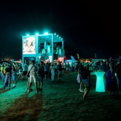Nočná festivalová scéna s osvetleným dvojpodlažným stánkom, okolo ktorého sa pohybujú skupiny návštevníkov; v popredí stojí vysoký svietiaci stolík na státie, v pozadí sú ďalšie stánky a tmavá obloha.