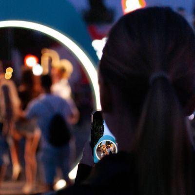 Osoba stojí v exteriéri a drží smartfón, ktorým fotografuje scénu pred sebou. V pozadí sú rozmazané postavy ľudí a svetelné IQOS logo, ktorý rámuje skupinu v diaľke. Scéna je v pozadí osvetlená farebnými umelými svetlami.
