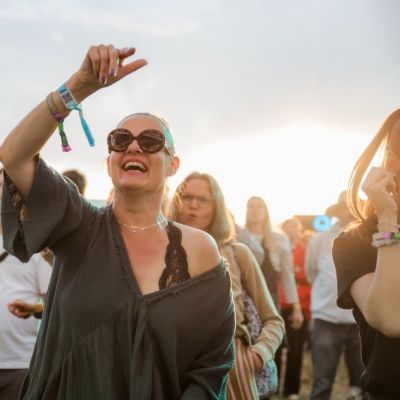 Osoba v slnečných okuliaroch s náramkami zdvíha ruku v dave ľudí na vonkajšom podujatí pri západe slnka.
