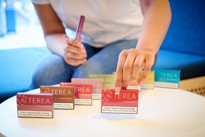 Eine TEREA Packung und ein TEREA Stick in den Händen eines Menschen vor dem Sortiment aller Geschmacksrichtungen
