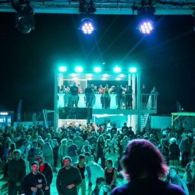 Dav ľudí stojí pred osvetleným pódiom a DJom na festivale v noci.