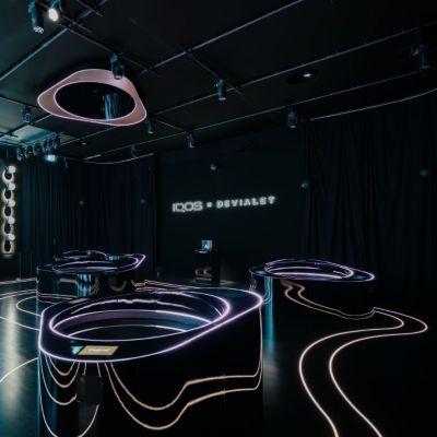 Interiér expozície IQOS × DEVIALET s podsvietenými výstavnými pultmi a logom na čiernej stene.