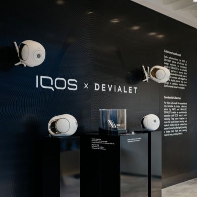 Reproduktory DEVIALET a produkt IQOS vystavené na podstavcoch pred stenou s logom IQOS × DEVIALET.