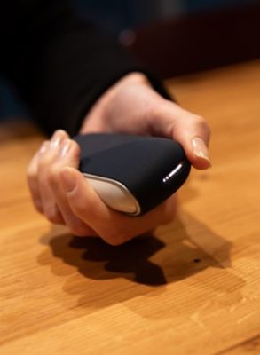 Eine Person hält ein eingeschaltetes IQOS ORIGINALS DUO Gerät mit leuchtender LED-Anzeige in der Hand.