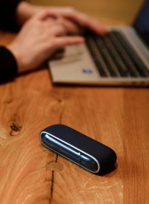 Ein blaues IQOS ORIGINALS DUO Gerät liegt auf einem Holztisch vor einer Person, die an einem Laptop arbeitet.