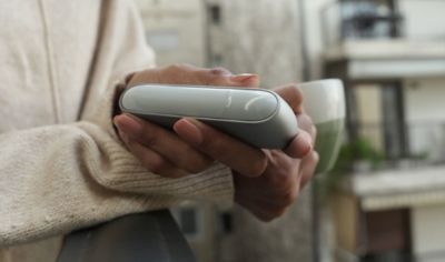 Person hält IQOS ORIGINALS DUO Silver in der einen und Tasse in der anderen Hand.