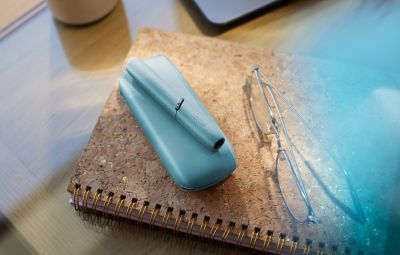 Appareil IQOS turquoise posé sur un carnet à spirales avec des lunettes à côté