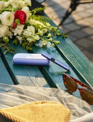 IQOS ILUMA i Digital Violet liegt auf eine Parkbank neben Blumen und Sonnenbrille