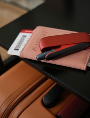 IQOS ORIGINALS DUO Scarlet liegt auf rosa Reisepass-Etui mit Flugticket auf schwarzem Tisch.