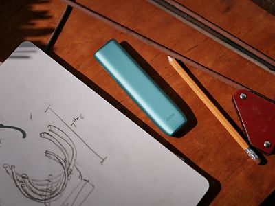 IQOS Iluma i ONE turquesa sobre mesa con herramientas y boceto técnico