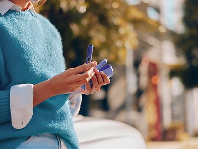 Persona sosteniendo IQOS Iluma i turquesa en exterior con fondo cálido