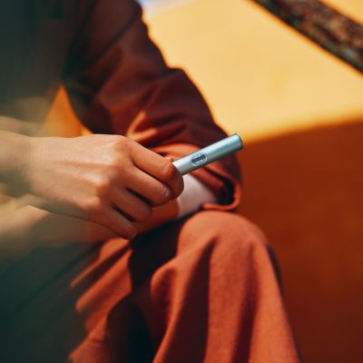 Mano sosteniendo un IQOS Iluma i turquesa en orientación vertical con fondo naranja.