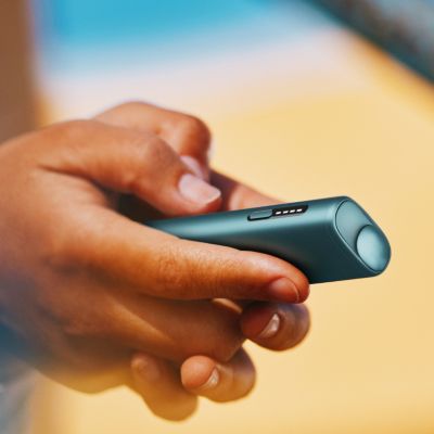 Mano sosteniendo un IQOS ILUMA ONE turquesa en un entorno soleado.