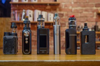 Six vape devices stand on a wooden counter, the background is blurred showing vapes on different shelves