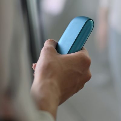 Person hält IQOS ORIGINALS DUO Turquoise in Hand vor unscharfem Hintergrund.