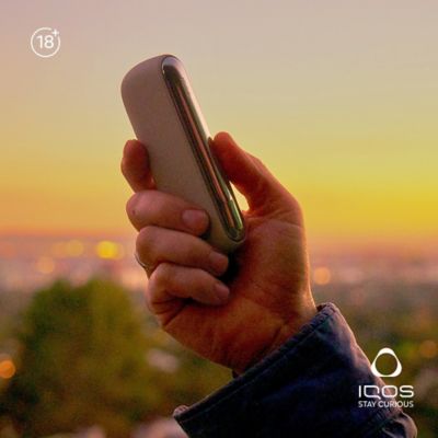 IQOS ILUMA Charger Pebble Beige held in hand against the sunset