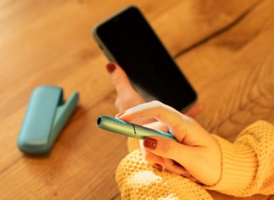 Person hält IQOS ORIGINALS DUO Turquoise Holder in rechter und Smartphone in linker Hand. Pocket Charger liegt im Hintergrund.