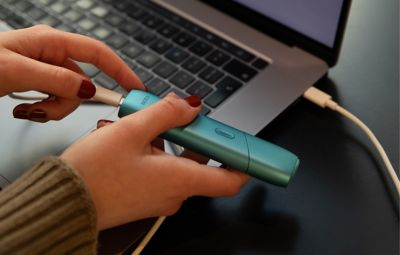 Person steckt IQOS ORIGINALS ONE Turquoise via USB-Kabel an Laptop an.