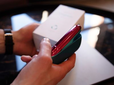 Person hält IQOS ORIGINALS DUO Turquoise mit rotem Klappen-Cover vor geöffneter Geräte-Verpackung.