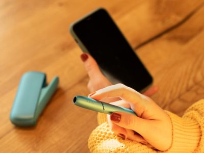 Frau hält ein IQOS ORIGINALS DUO Turquoise in der rechten Hand  und ein Smartphone in der linken.