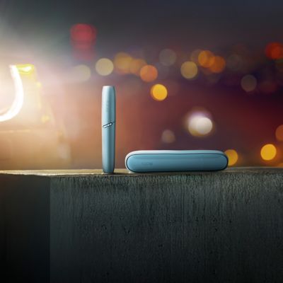 Ein blaues IQOS ORIGINALS DUO Gerät liegt auf einer Betonoberfläche vor verschwommenen Stadtlichtern bei Nacht.